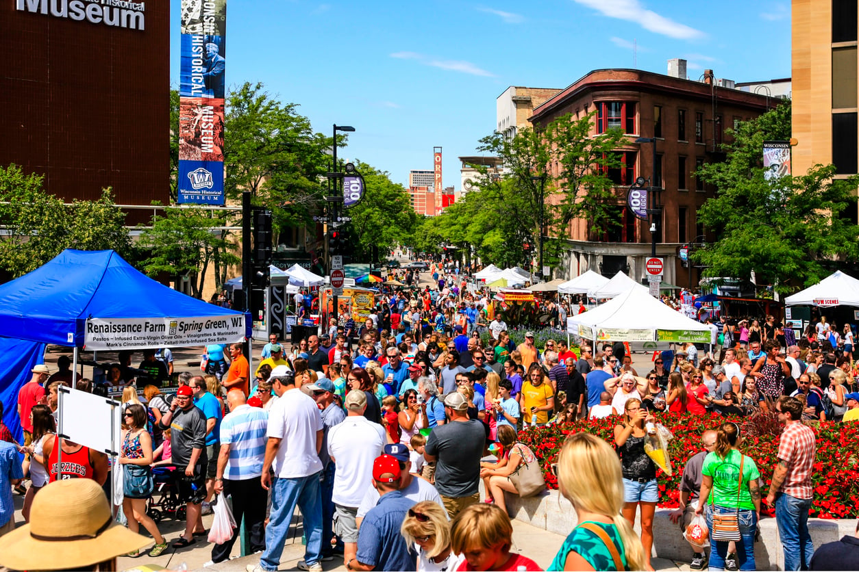A crowded outdoor street festival with white vendor tents, a blue canopy for Renaissance Farm, and people walking along a city street lined with brick buildings.