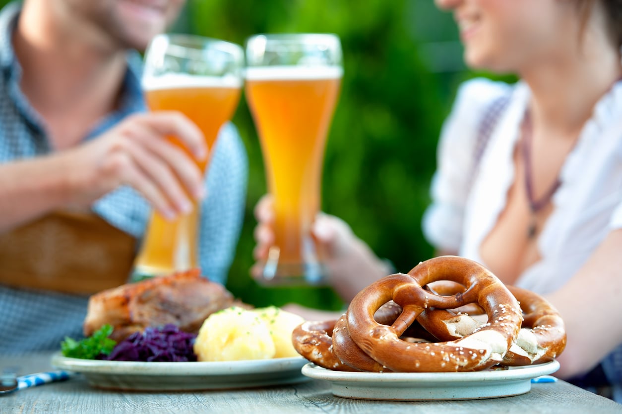Two people toasting with tall glasses of beer, with plates of pretzels and German food on the table in front of them.
