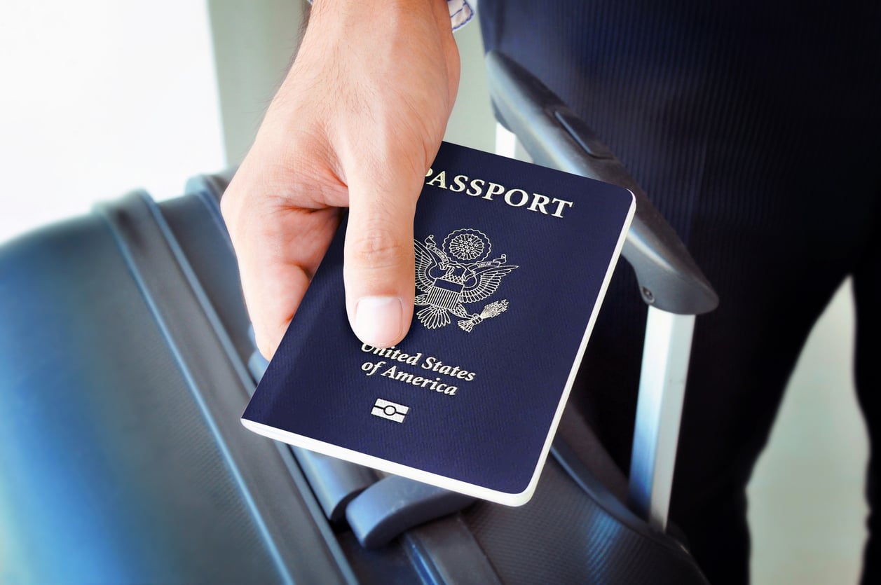 vacation expenses A person holding a United States passport in front of a piece of rolling luggage.