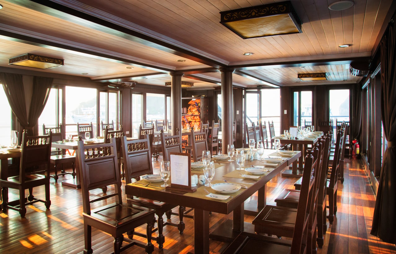 The dining room of a boat featuring rows of wooden tables and chairs set for service with glassware and plates.