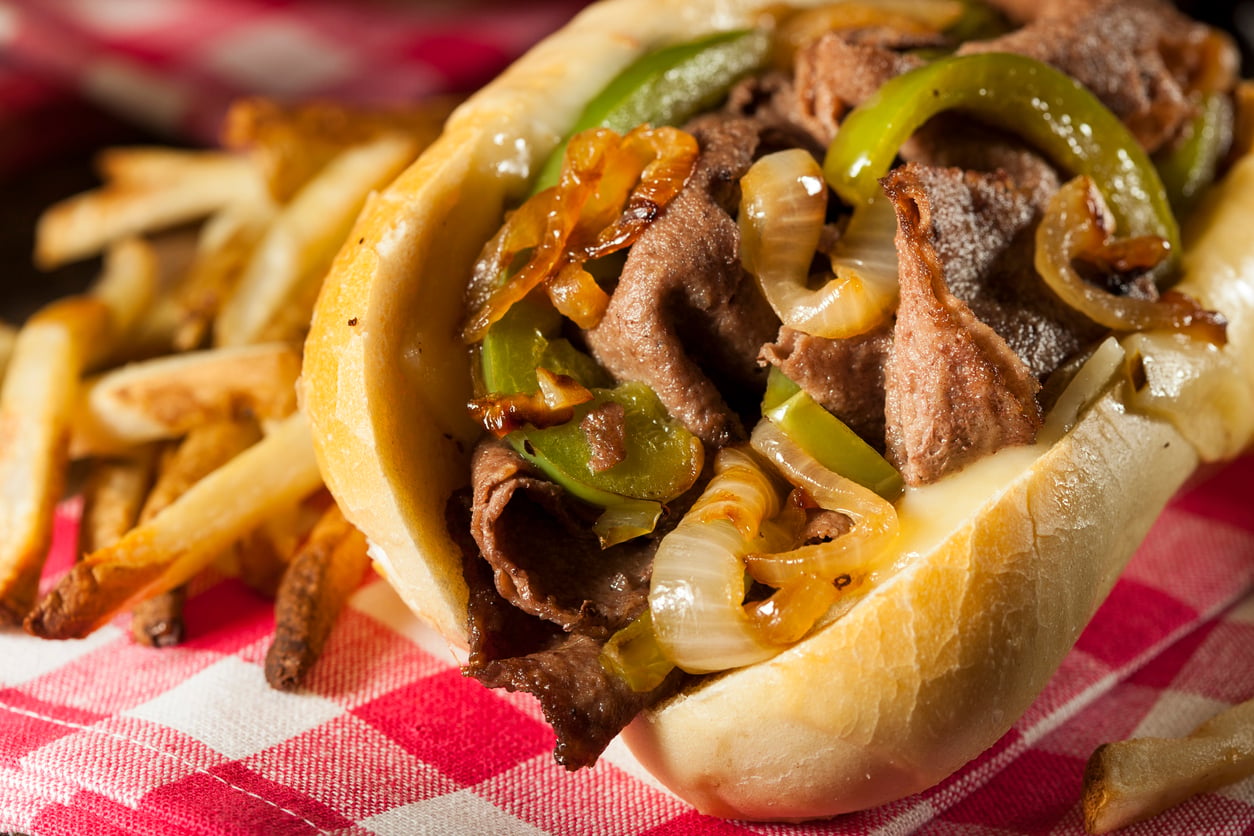 A cheesesteak sandwich with sautéed peppers and onions, served with a side of french fries on a red and white checkered napkin.