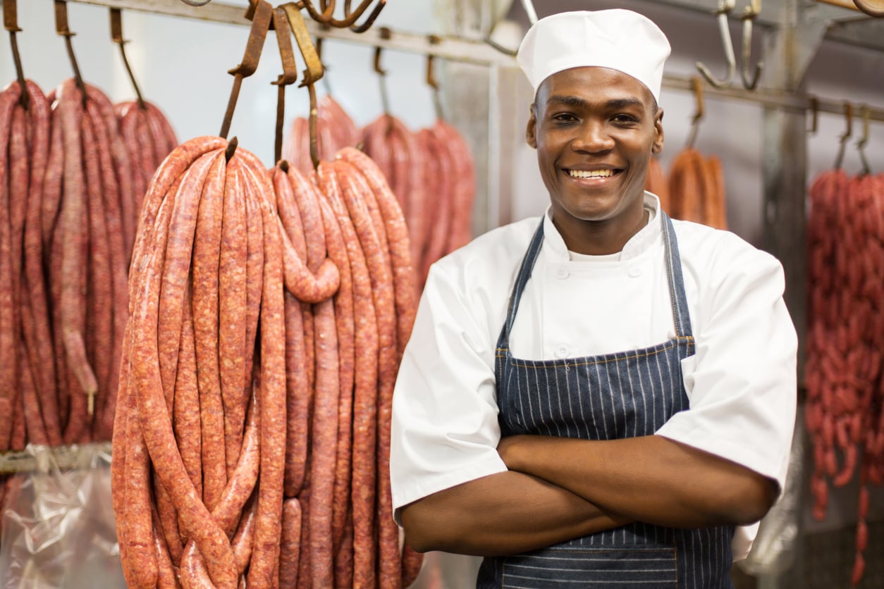 A smiling butcher in a white uniform and apron stands with his arms crossed in front of rows of hanging sausages.