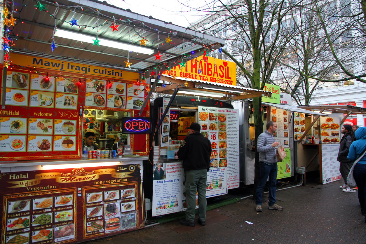 Three side-by-side food carts in an outdoor market, featuring menus for Iraqi cuisine, Thai food, and Bento Korean BBQ, with a few customers waiting to order.