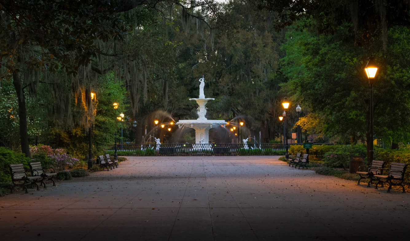 A multi-tiered white fountain in a park at dusk, surrounded by trees with Spanish moss, lampposts, and wooden benches along a paved path.