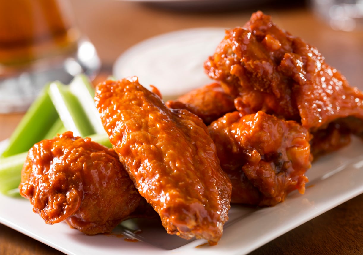 A pile of buffalo wings on a white plate with celery sticks and a side of dip.