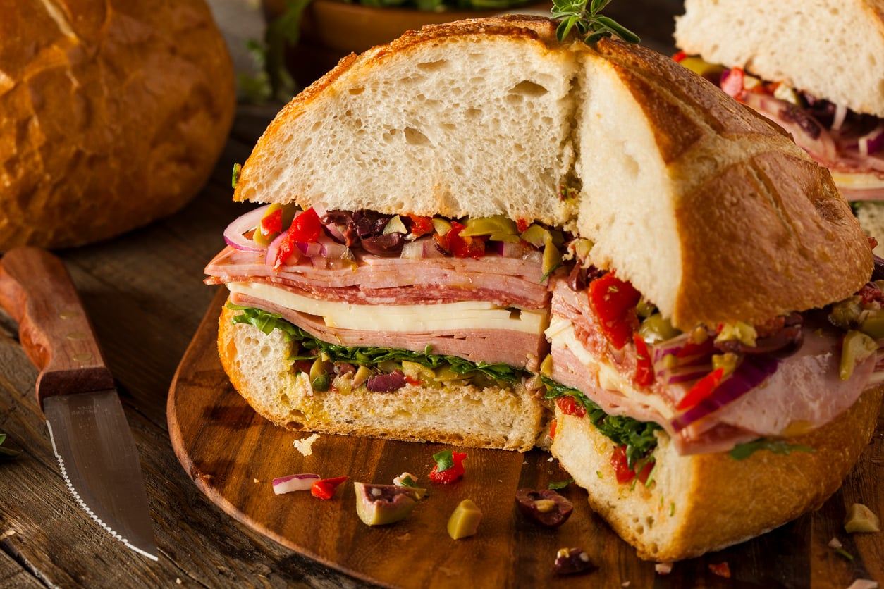 A round muffuletta sandwich sliced into quarters, layered with cured meats, cheese, and olive salad on a wooden board with a knife nearby.