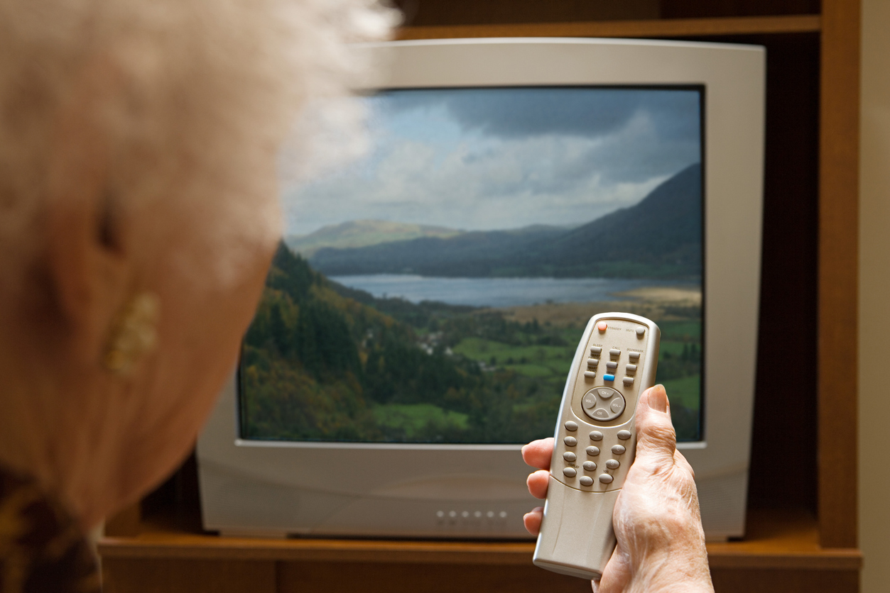 An older person holding a remote control while watching a television screen that displays a landscape of mountains and a lake.