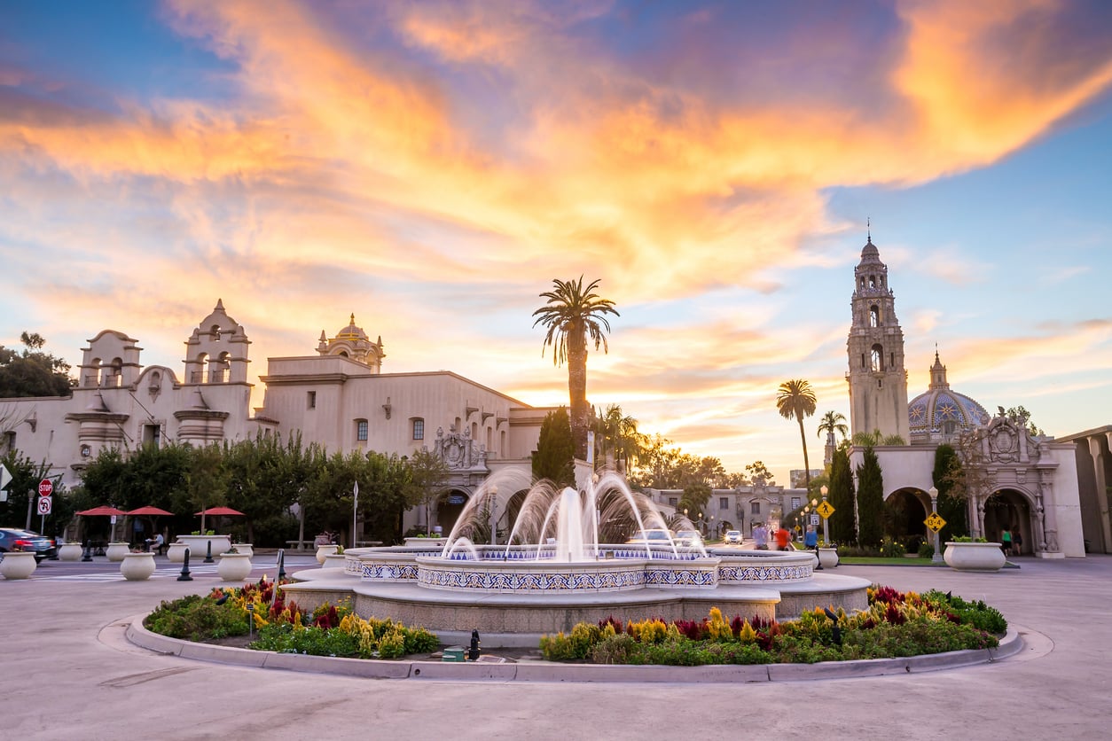 A decorative fountain with colorful flowers sits before historic Spanish-style buildings and a tall tower at sunset—perfect for vacations where the hotel barely matters. Dramatic clouds fill the sky above.