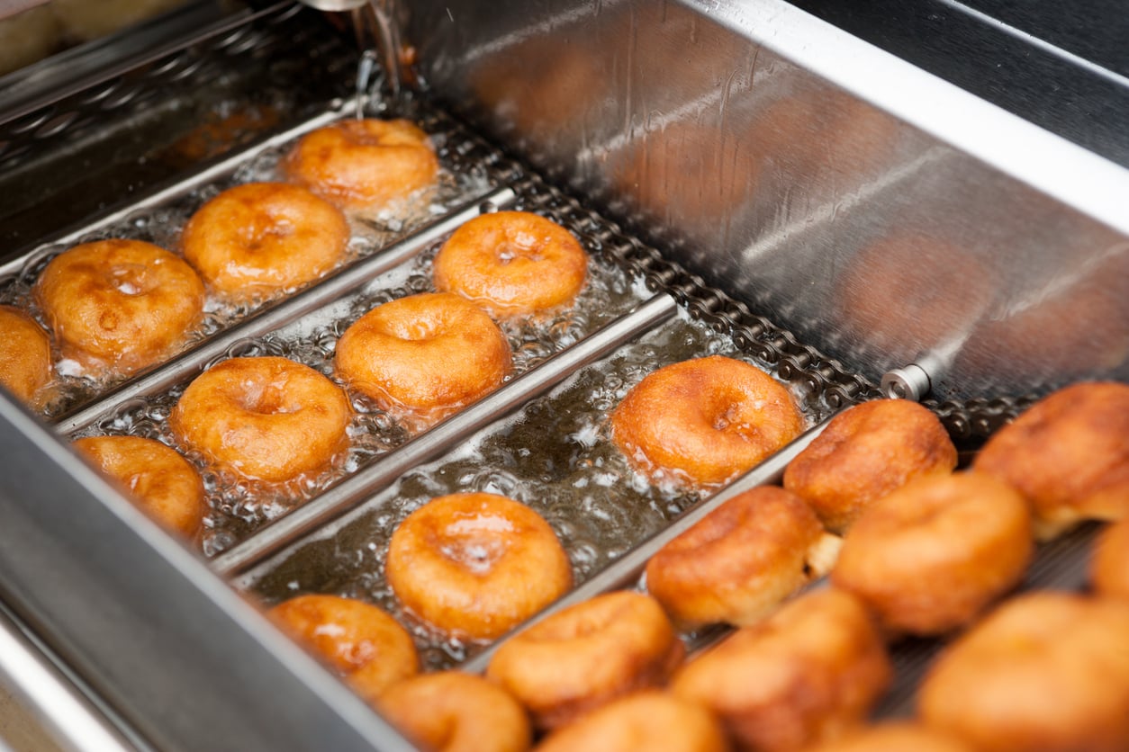 Donuts frying on a conveyor belt in a commercial deep fryer.