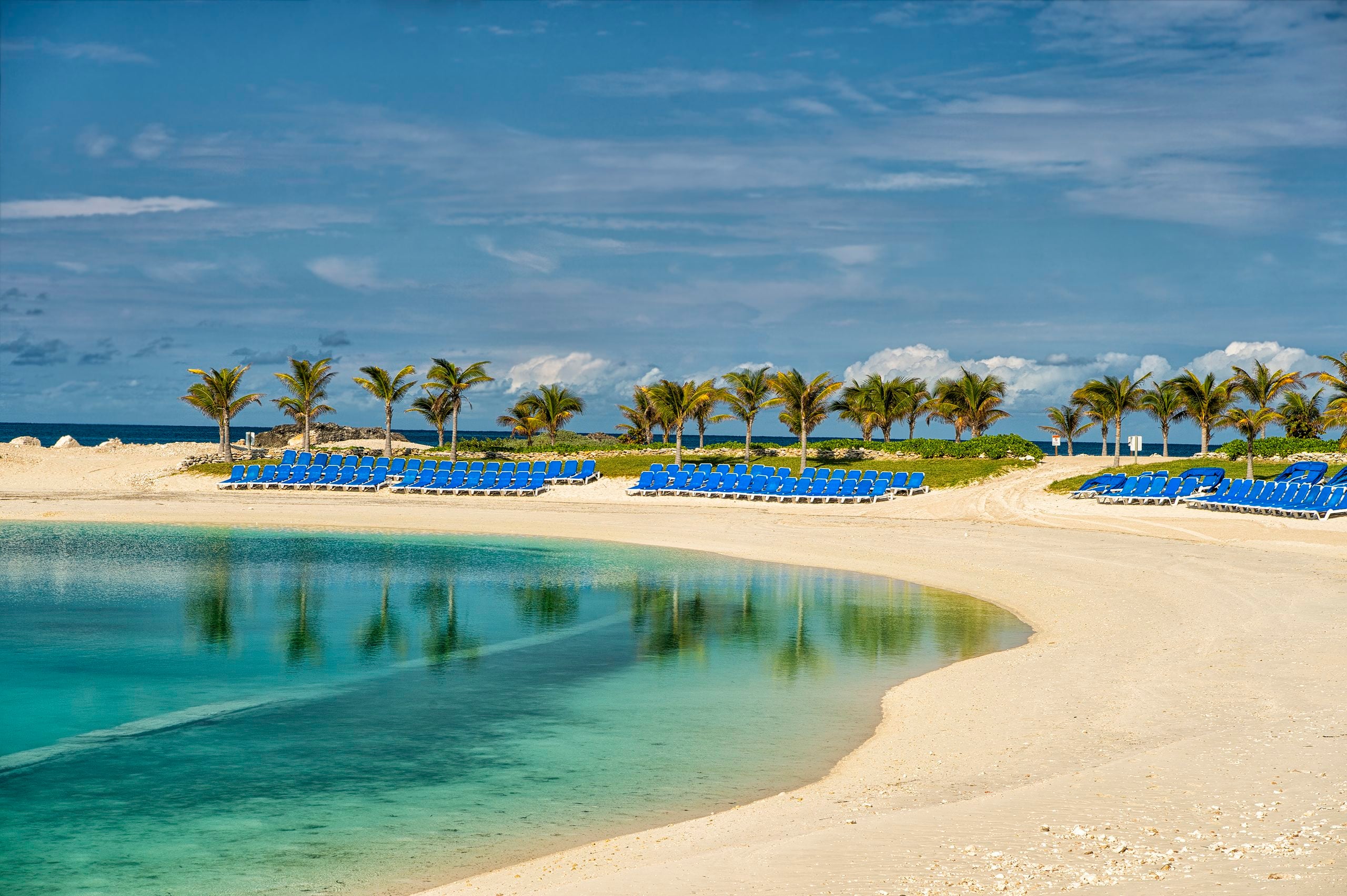 Rows of blue lounge chairs on a sandy beach bordering a turquoise lagoon, with a line of palm trees under a blue sky.