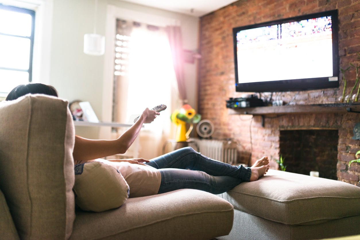 A person reclining on a sofa holds a remote control toward a television mounted on a brick wall.