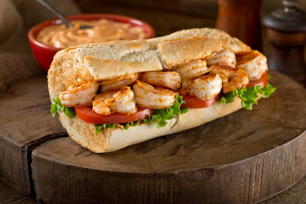 Shrimp po’ boy sandwich with lettuce and tomato on a wooden serving board, with a small bowl of sauce in the background.