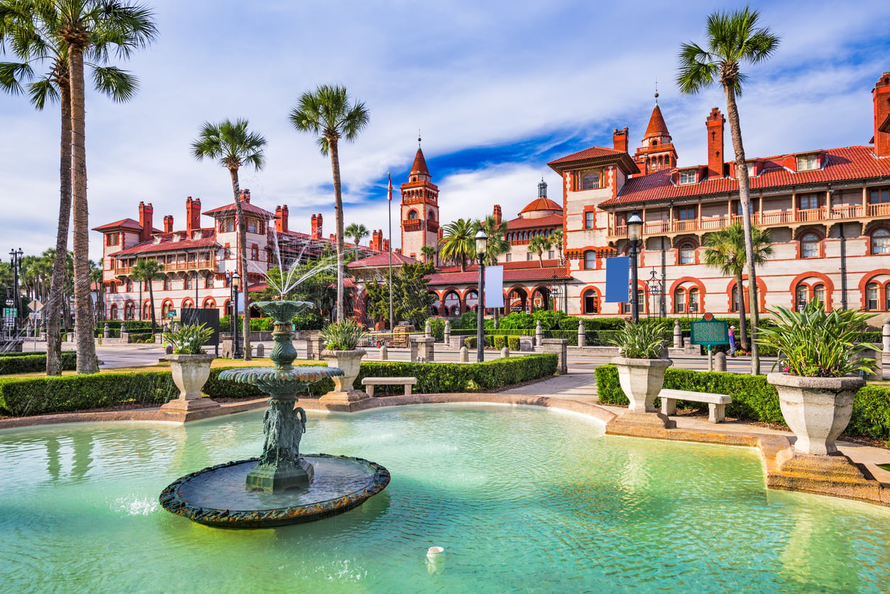 A historic Spanish-style building with red roofs stands behind a fountain and palm trees, offering the grandeur of American Destinations That Feel Bigger, all under a blue sky and surrounded by manicured gardens.