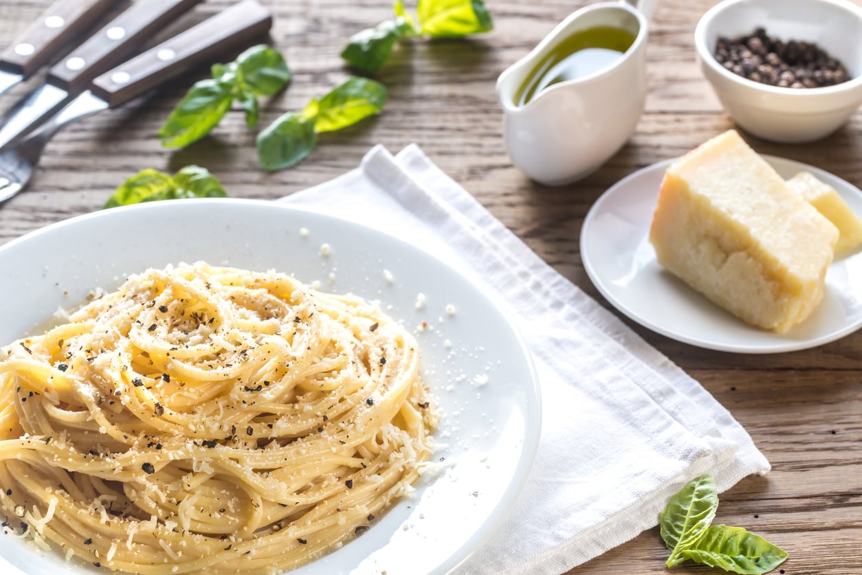 A plate of spaghetti cacio e pepe garnished with grated cheese and black pepper, served with a wedge of parmesan, olive oil, and fresh basil.