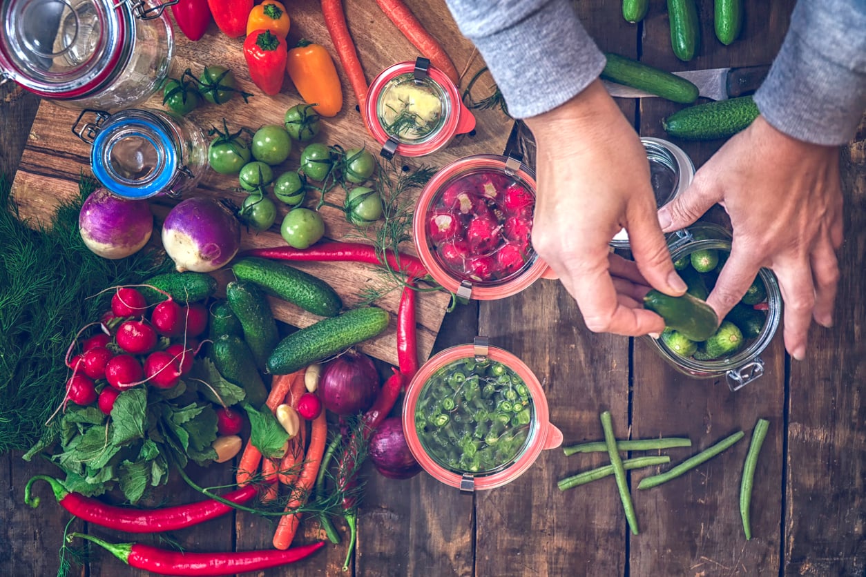 A person’s hands pack small cucumbers into a glass canning jar, surrounded by various fresh vegetables and open jars on a wooden table.