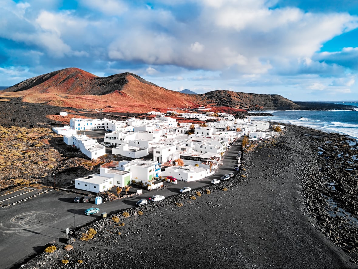 Aerial view of a white village with a road running along a black volcanic beach beneath red mountains.