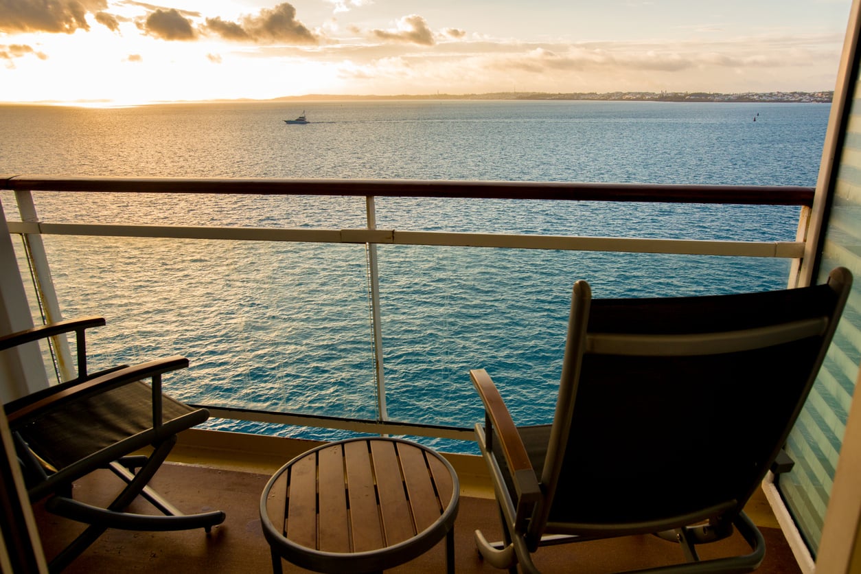 View of the ocean from a cruise ship balcony featuring two chairs, a small table, and a distant boat at sunset.