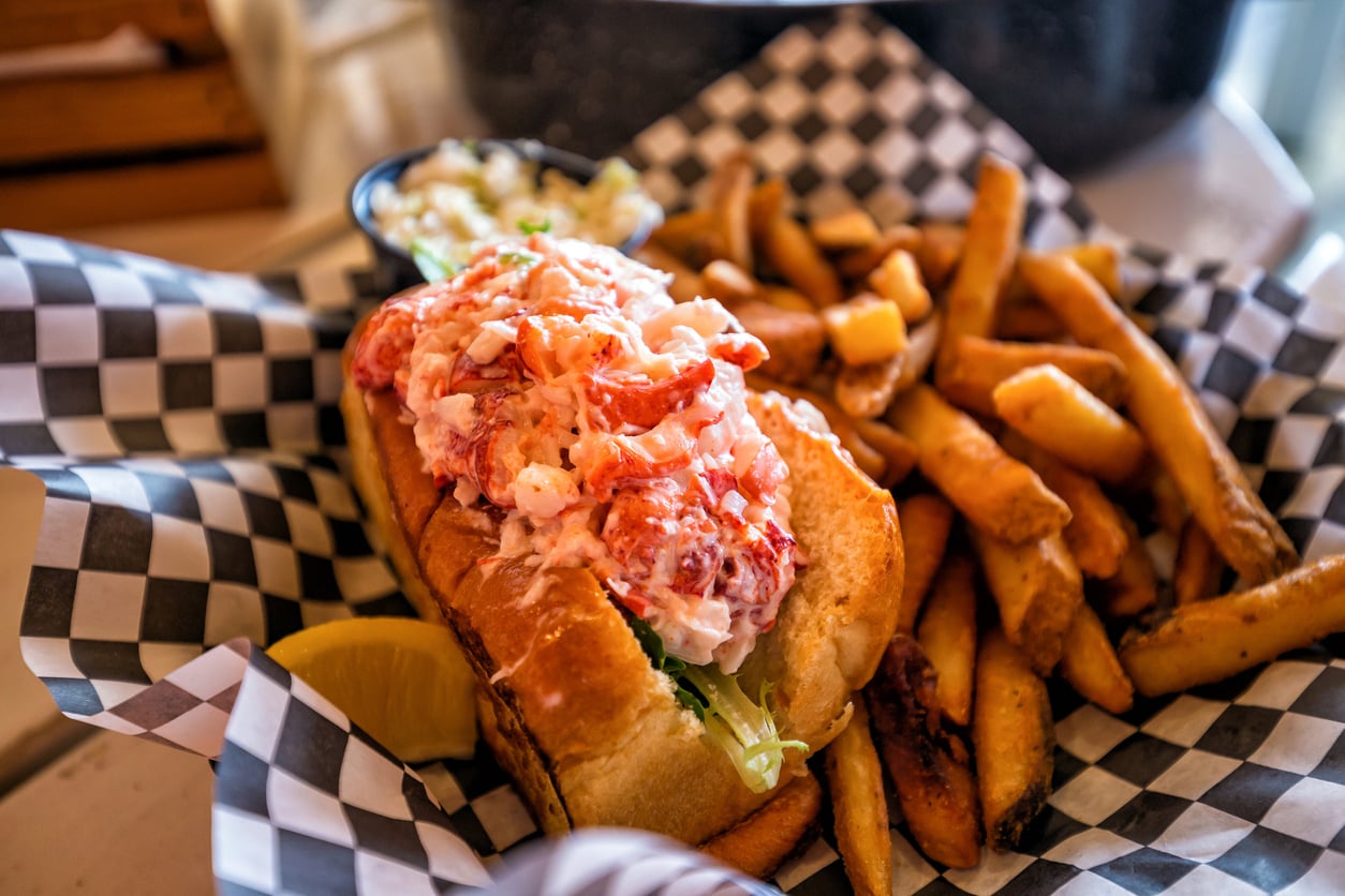 A lobster roll served with a side of french fries and coleslaw in a basket lined with checkered paper.