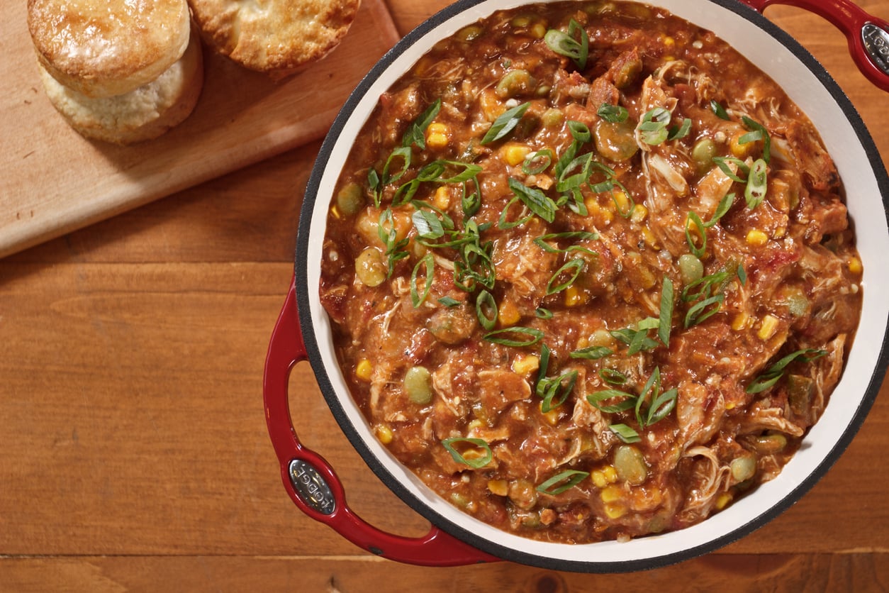 A red Dutch oven filled with chicken and vegetable stew topped with sliced green onions, sitting on a wooden surface next to two biscuits on a cutting board.