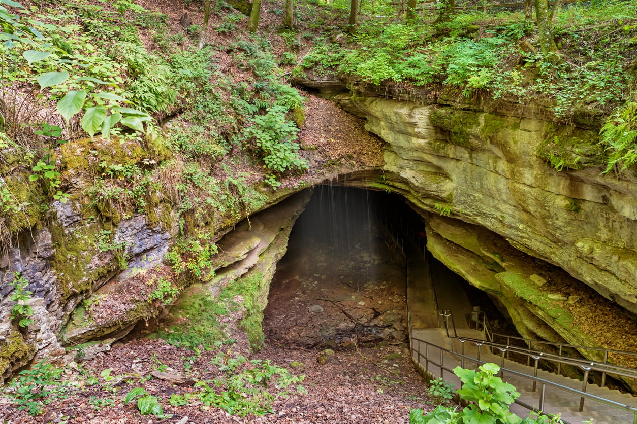 A stone staircase leads down into a wide cave entrance with a small waterfall trickling over the ledge.