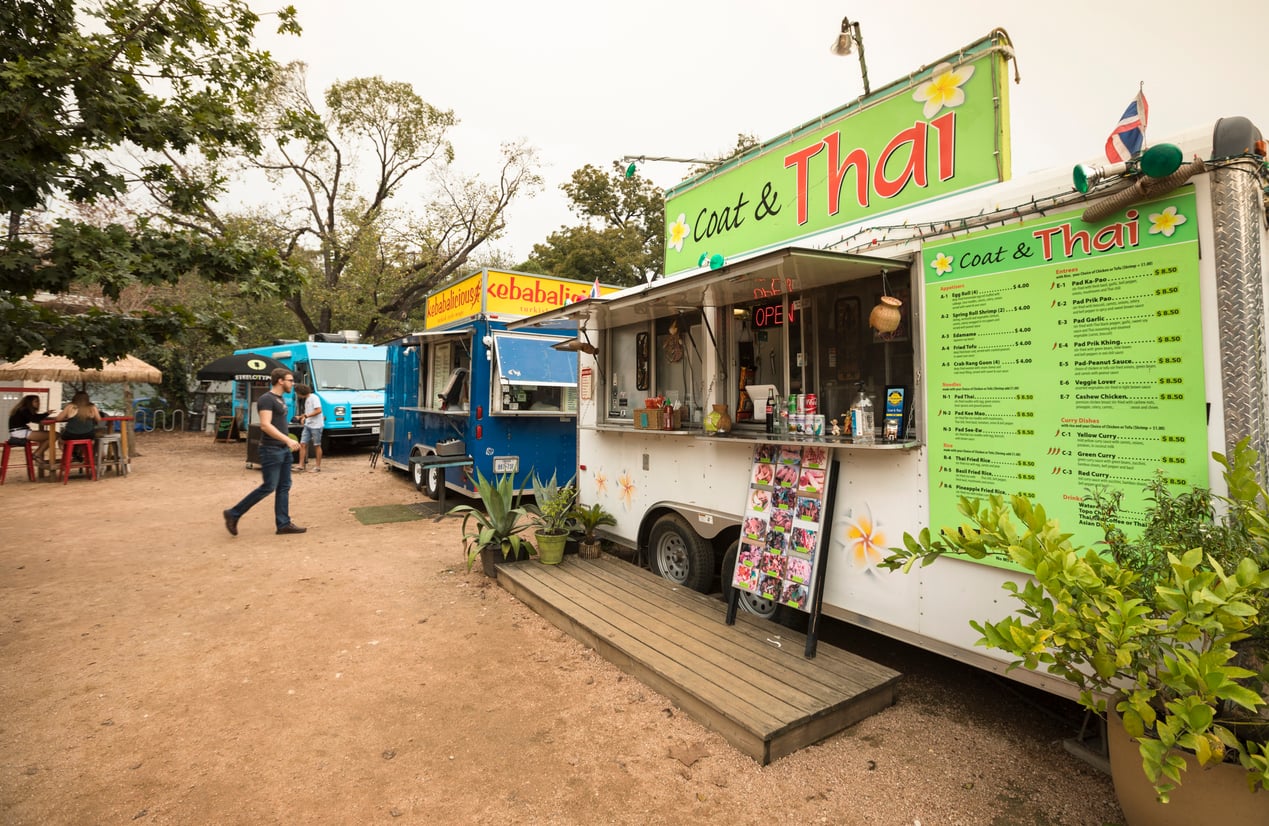 A Coat & Thai food truck with a menu board parked next to other food trucks in a dirt lot.