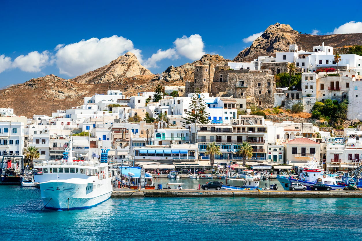 A white ferry docked in a harbor in front of a hillside town of white buildings and a stone fortress under a blue sky.