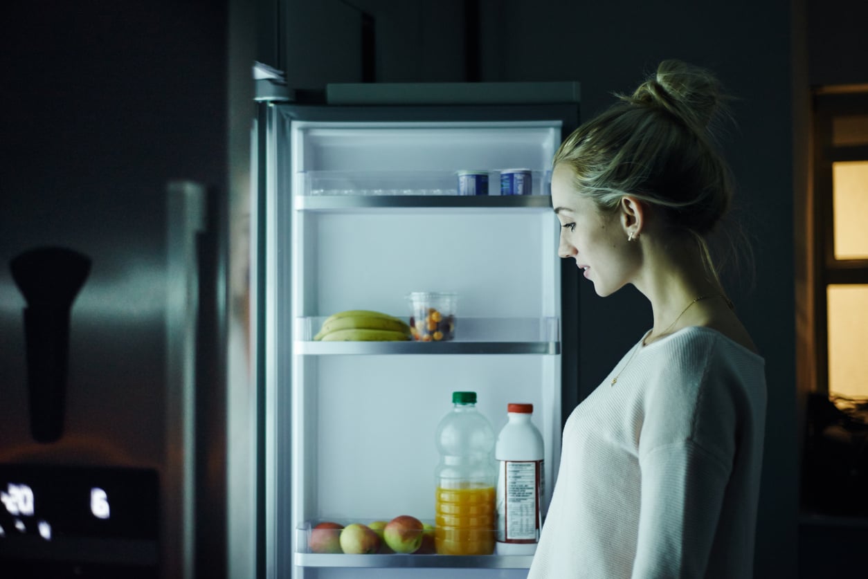 A young woman stands in a dark kitchen, looking into the illuminated interior of an open refrigerator.