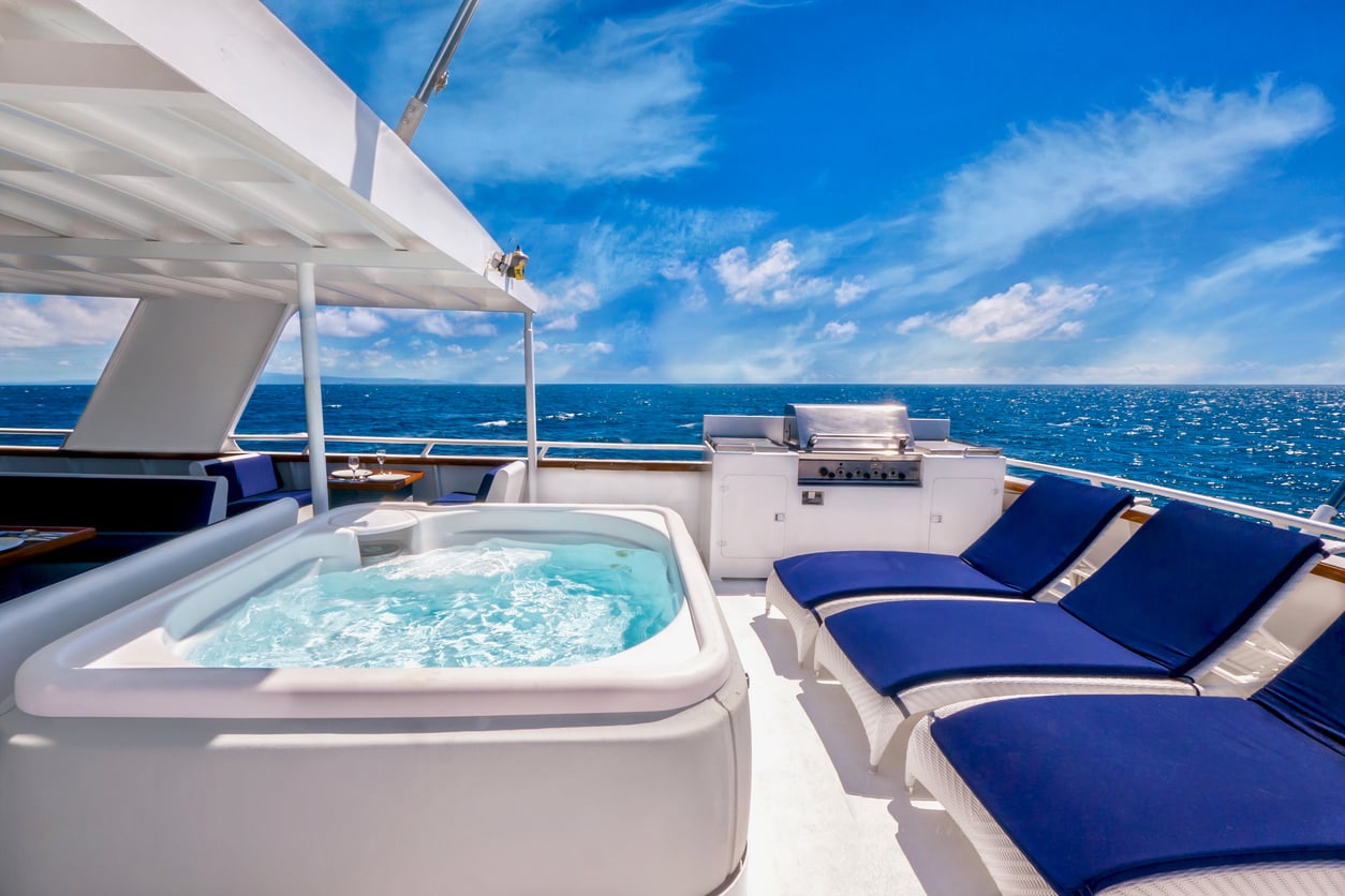 A white hot tub, a stainless steel grill, and three blue lounge chairs on the deck of a yacht overlooking the ocean, reflecting luxury cruise ship charges.