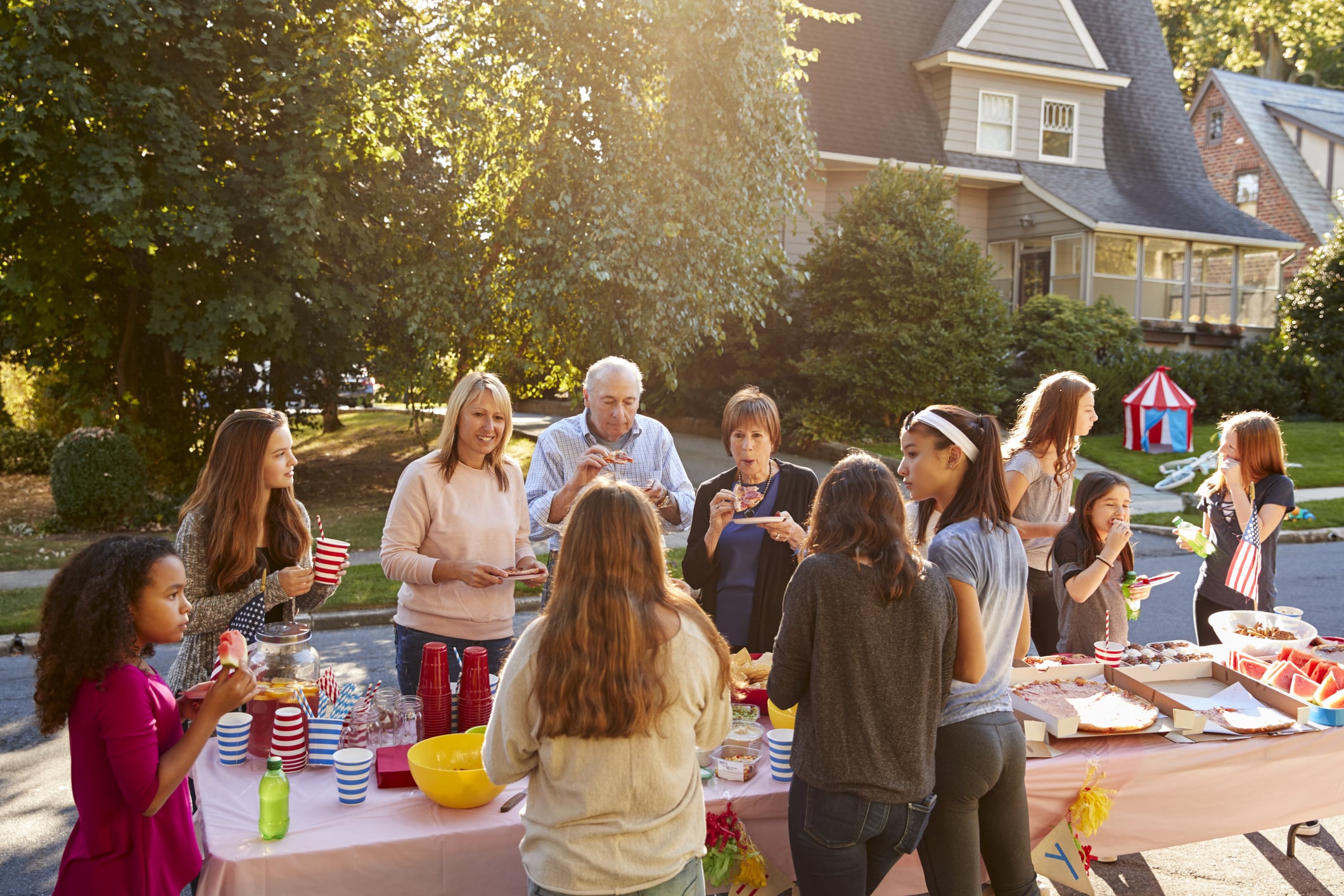 Things Americans are doing less often: A diverse group of people of various ages gather around a table set up outdoors with food and drinks for a celebration.
