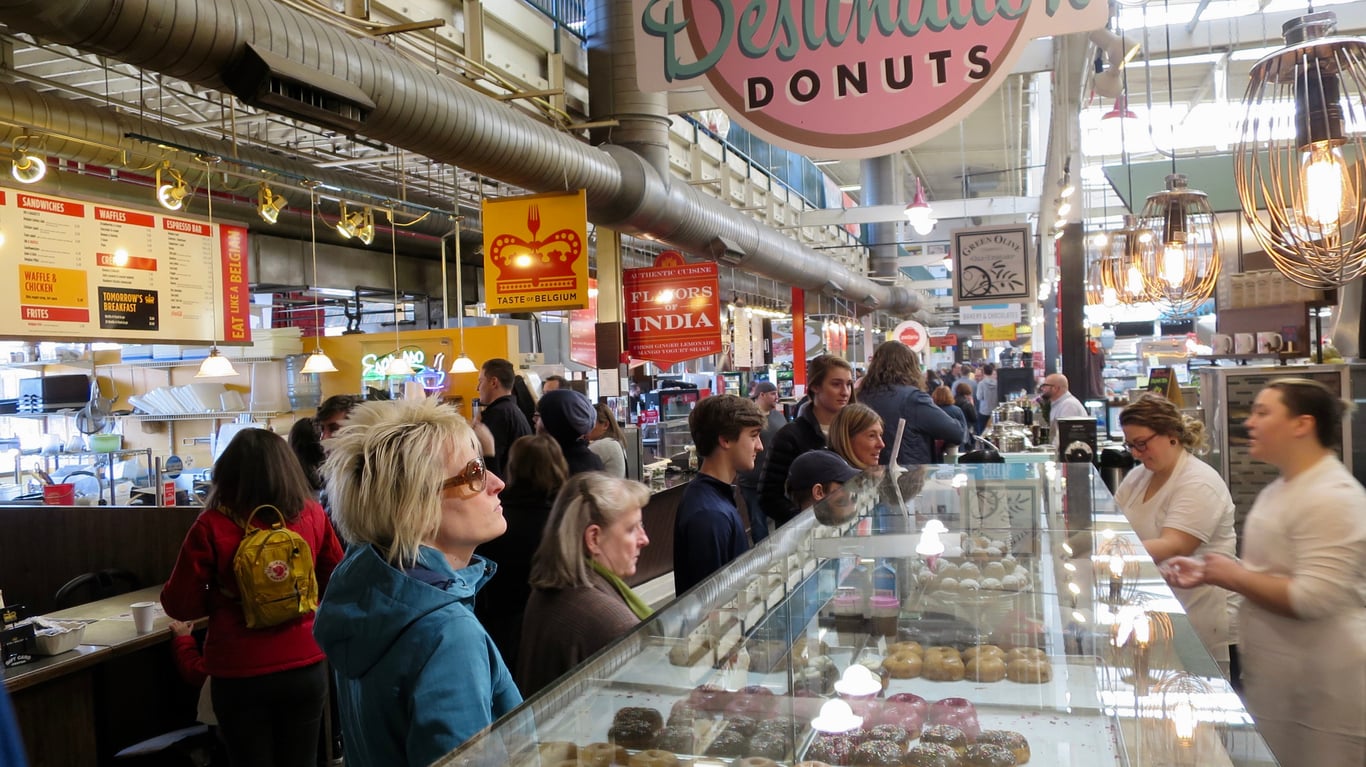 Customers wait in line at a food hall in front of a glass display case filled with donuts, with “Destination Donuts” signage hanging above.