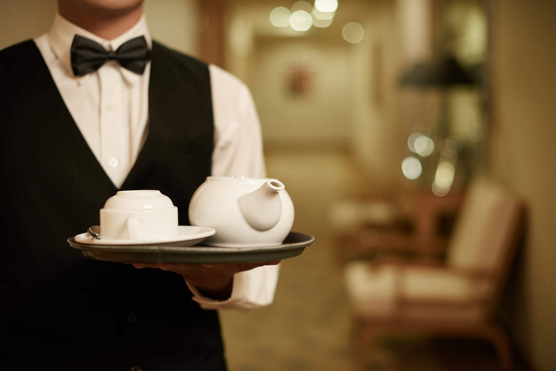 A server in a white shirt and black vest holding a tray with a teapot and teacup.