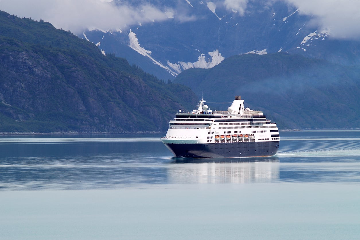 A large cruise ship sails through calm blue water, with tree-covered hills and snowy mountains in the background under a cloudy sky.