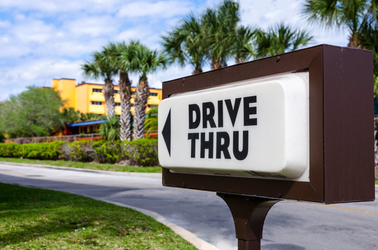 A drive-thru sign with a left-pointing arrow, positioned next to a road with palm trees and a yellow building in the background.