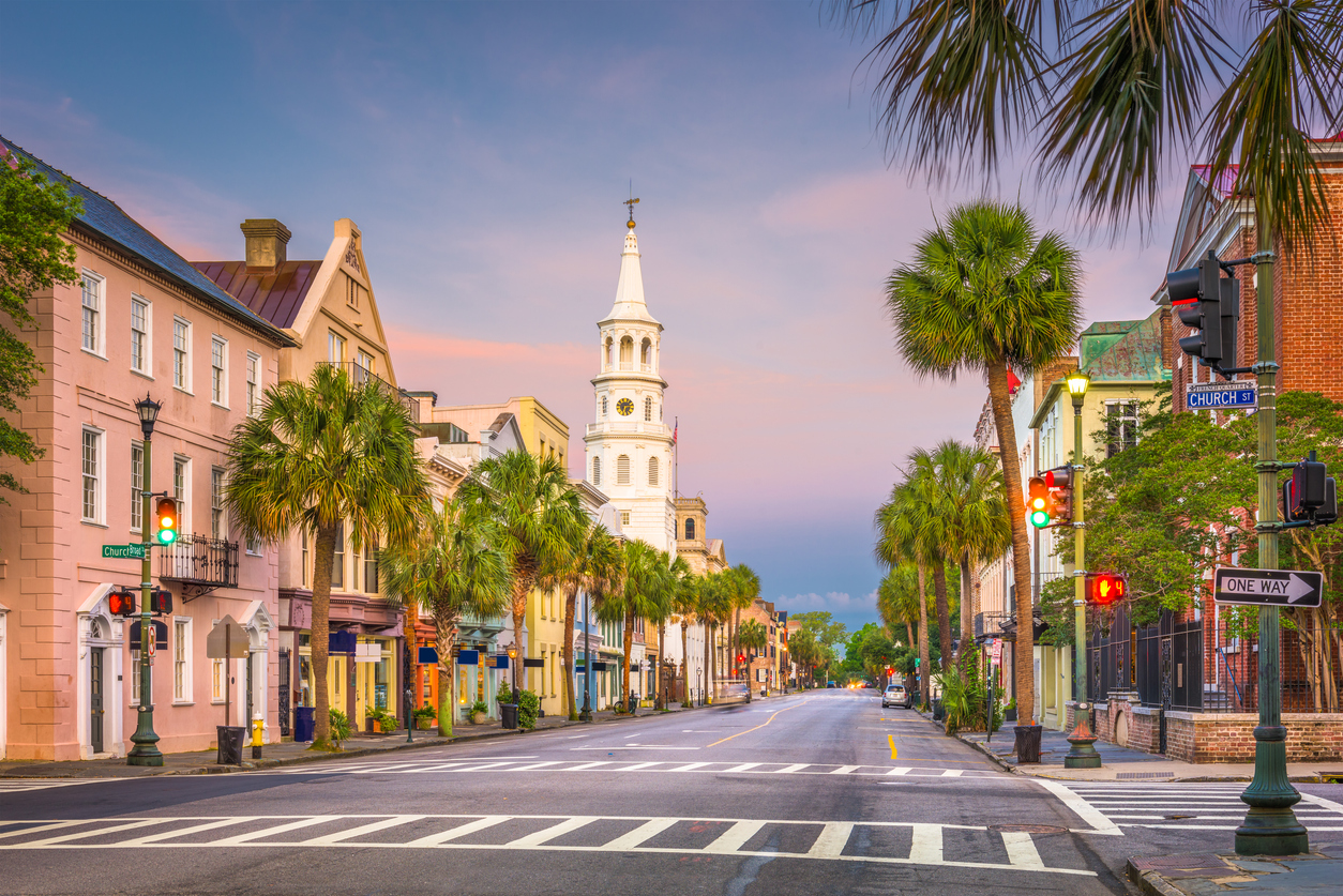 A street view of historic Charleston, South Carolina, featuring palm trees, multi-colored buildings, and the white steeple of St. Philip’s Church under a sunset sky during trips that feel luxurious.