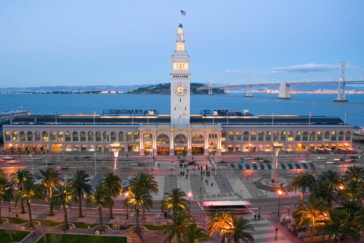 The Ferry Building in San Francisco with its clock tower at twilight, with the Bay Bridge visible in the distance.