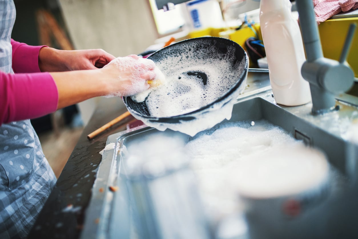 A person in an apron washes a soapy frying pan in a kitchen sink.