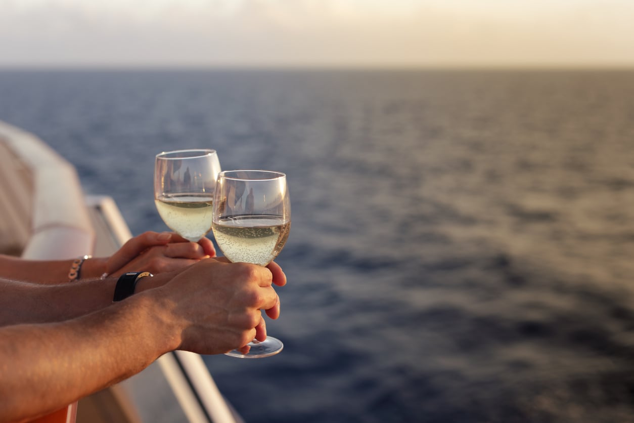 Two people holding glasses of white wine on a cruise ship deck overlooking the ocean at sunset.