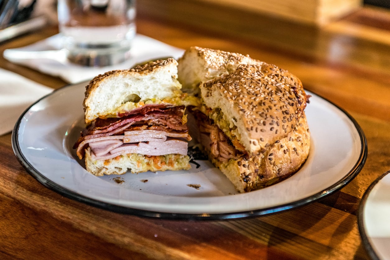 A sliced sandwich with layers of ham and bacon on a seeded bun, served on a white plate.