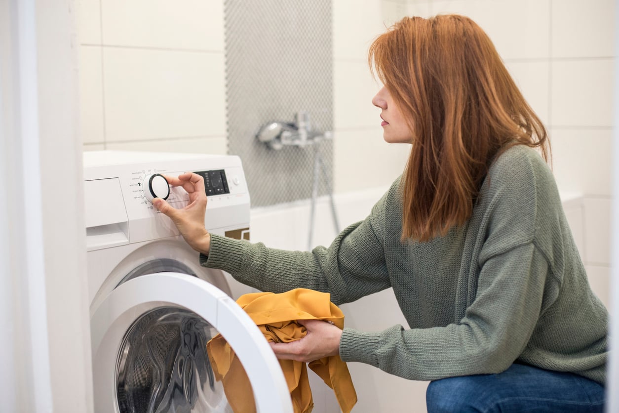 A person kneels to adjust the dial on a white washing machine while holding a yellow garment.