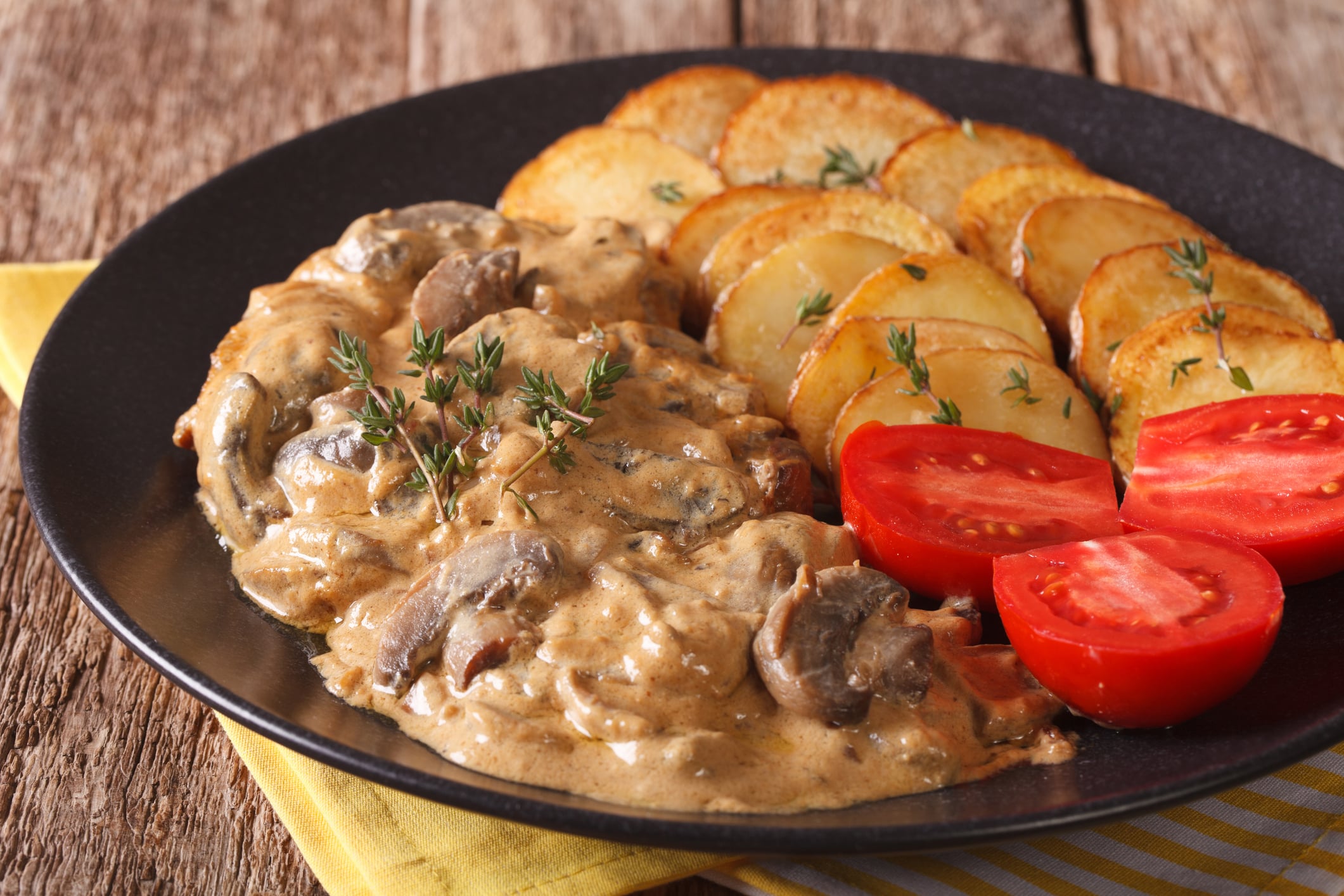 Meat with creamy mushroom sauce, served with roasted potato slices and halved cherry tomatoes on a dark plate.