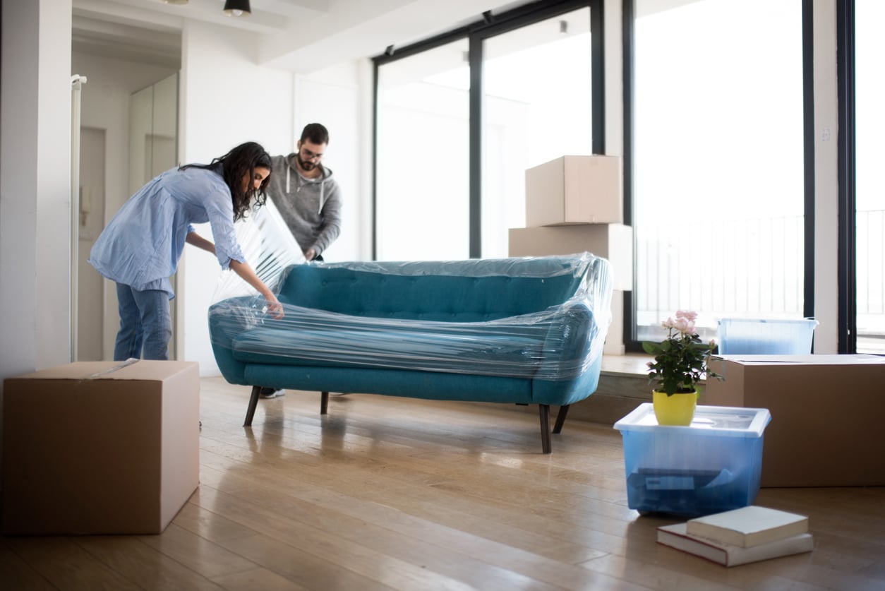 A man and woman wrap a blue sofa in plastic film in a room filled with cardboard moving boxes.