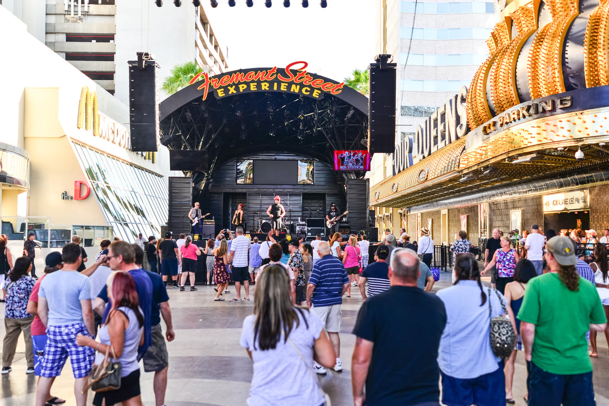 A band performs on a stage under the Fremont Street Experience canopy, with a crowd of people watching in the pedestrian mall.