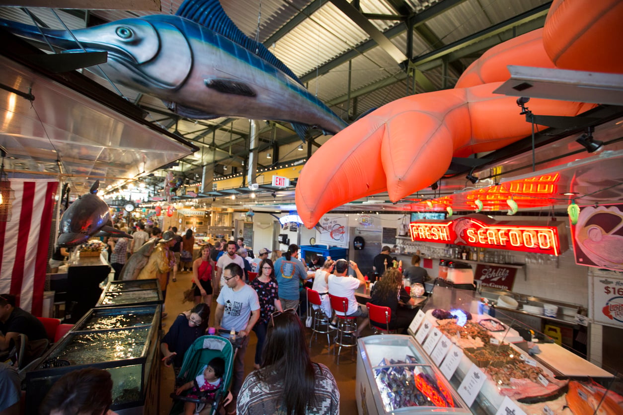 Indoor seafood market with a large model fish and lobster hanging from the ceiling above a counter with a red “FRESH SEAFOOD” neon sign and glass display cases filled with fish.