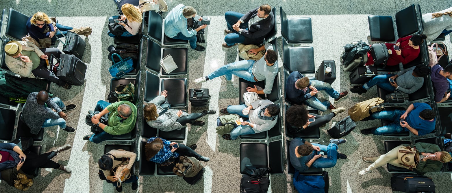An overhead view of passengers sitting in airport terminal chairs, many checking their mobile devices to make trip planning decisions with luggage placed at their feet.