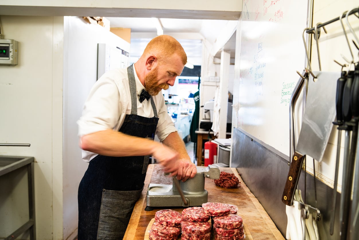 A butcher in a white shirt, bow tie, and apron uses a press to form ground meat patties on a wooden counter.