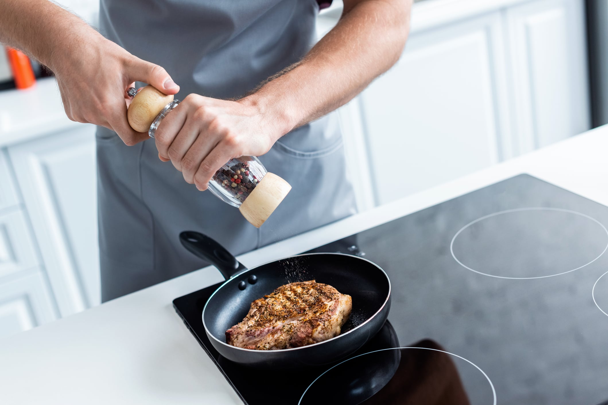 A person in a gray apron uses a pepper mill to season a seared steak in a frying pan on an electric stovetop.