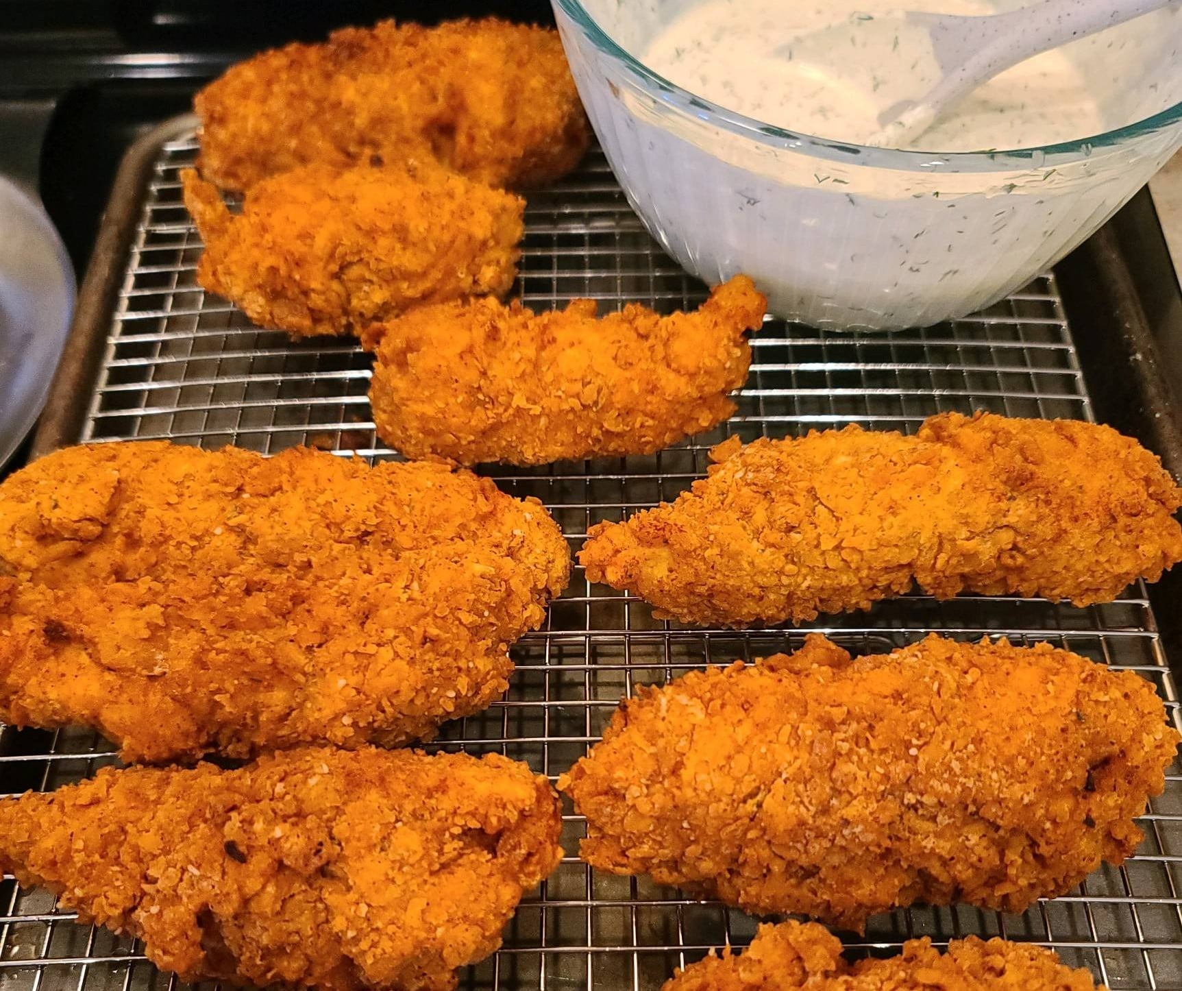 Several pieces of breaded, golden-brown fried chicken on a wire cooling rack next to a bowl of creamy herb dipping sauce.