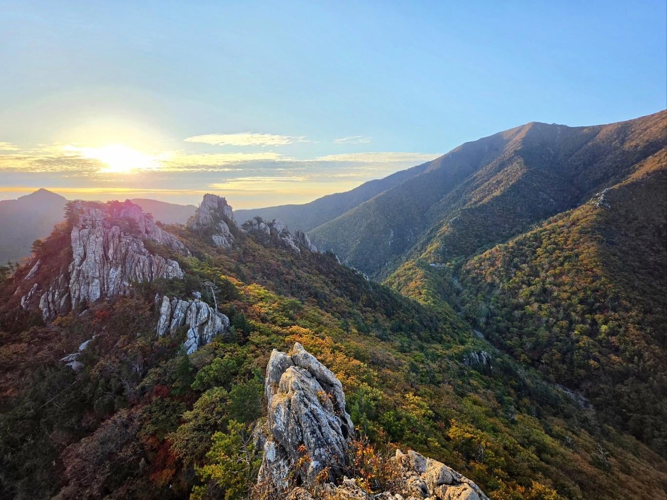 The sun rises over a jagged mountain ridge covered in autumn trees.