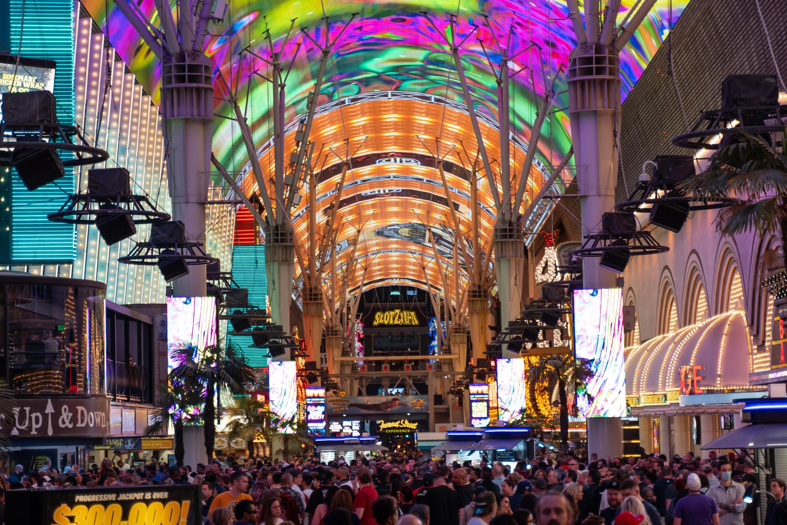 A large, dense crowd gathers beneath the massive, brightly lit LED canopy of the Fremont Street Experience in Las Vegas.