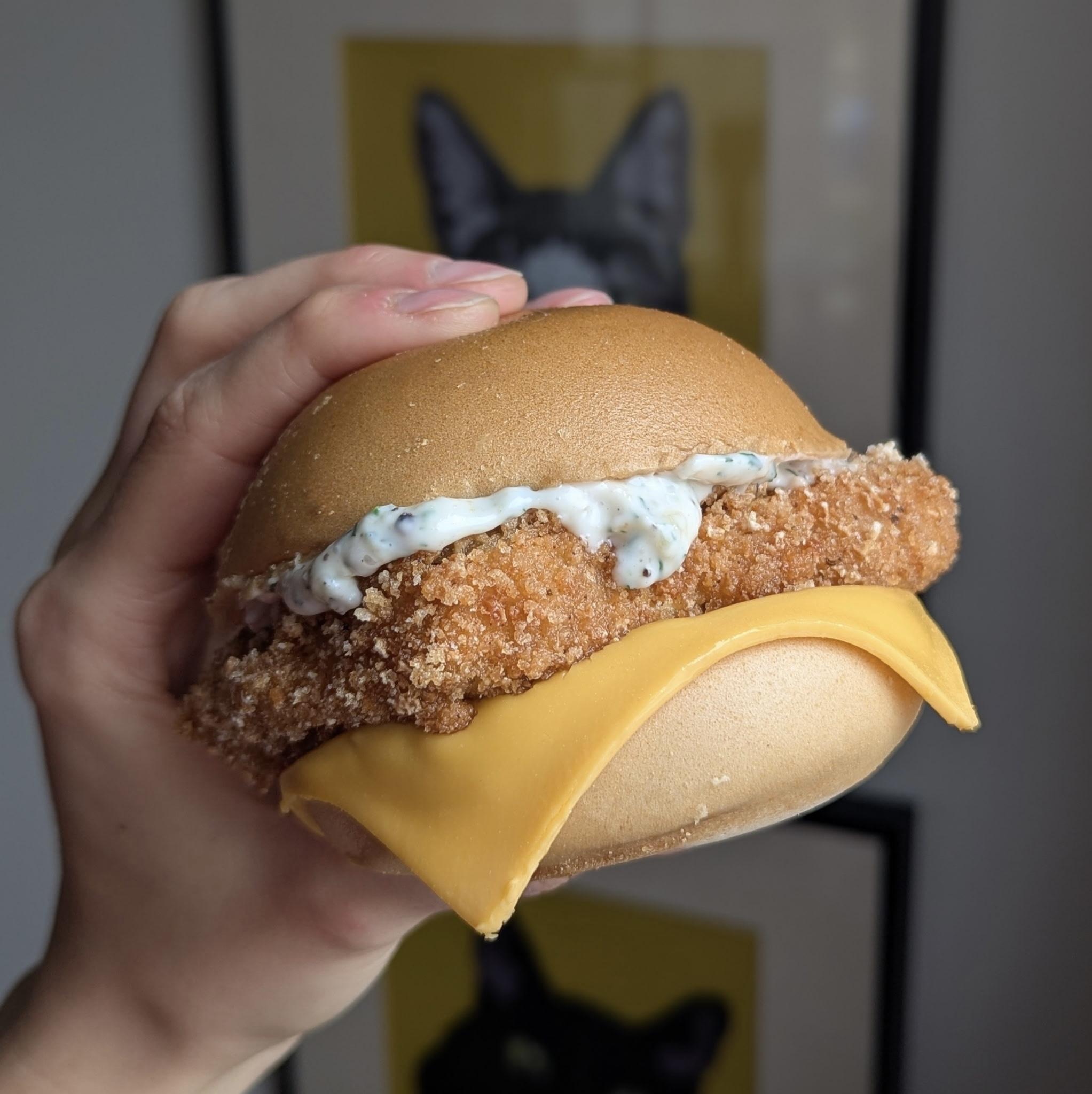 A hand holds a crispy fried chicken sandwich with melted cheese and creamy sauce on a round bun.